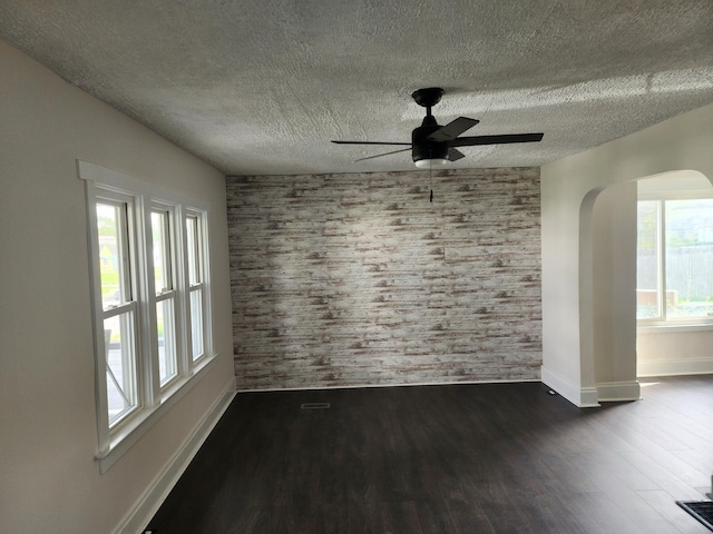 empty room featuring a textured ceiling, baseboards, ceiling fan, arched walkways, and dark wood-style floors