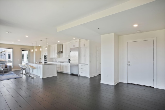 kitchen featuring stainless steel fridge, open floor plan, wall chimney range hood, a kitchen island with sink, and white cabinets