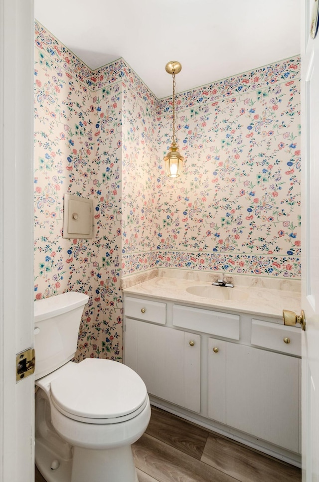 half bathroom with toilet, vanity, wallpapered walls, and wood finished floors