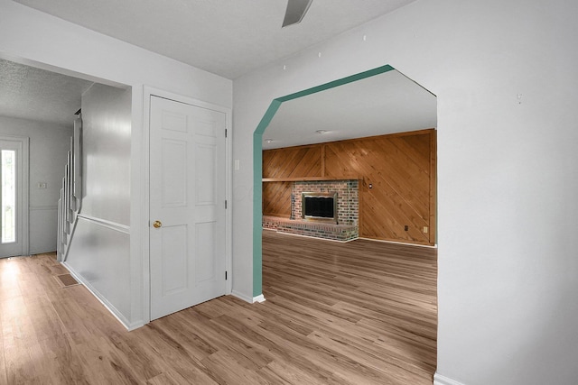unfurnished living room featuring wooden walls, a brick fireplace, a ceiling fan, baseboards, and light wood-style floors