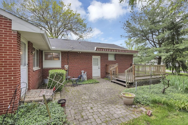 back of property with a deck, brick siding, a patio area, and roof with shingles