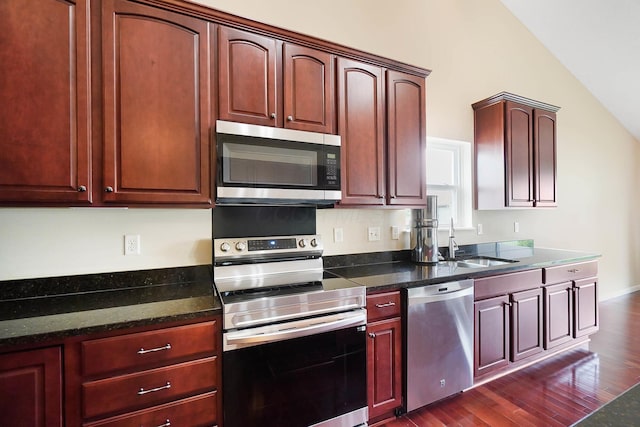 kitchen featuring appliances with stainless steel finishes, a sink, dark wood-style floors, lofted ceiling, and dark stone counters