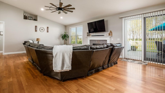 living area with a ceiling fan, light wood finished floors, a tile fireplace, vaulted ceiling, and baseboards