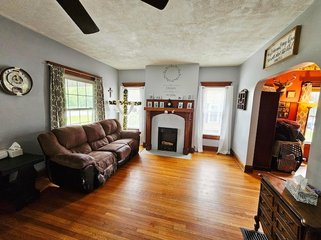 living area with ceiling fan, light wood-style flooring, a fireplace with flush hearth, arched walkways, and a textured ceiling