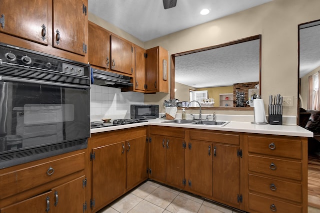 kitchen featuring black appliances, a sink, under cabinet range hood, brown cabinets, and tasteful backsplash