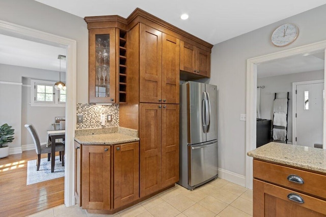 kitchen featuring freestanding refrigerator, brown cabinetry, baseboards, glass insert cabinets, and light stone countertops