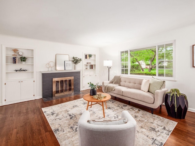 living area with built in shelves, dark wood-type flooring, a brick fireplace, baseboards, and crown molding