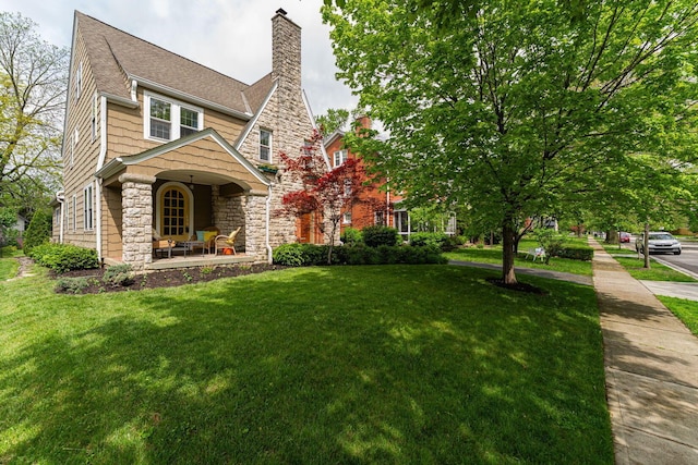 view of front of property with covered porch, stone siding, a front yard, a chimney, and roof with shingles