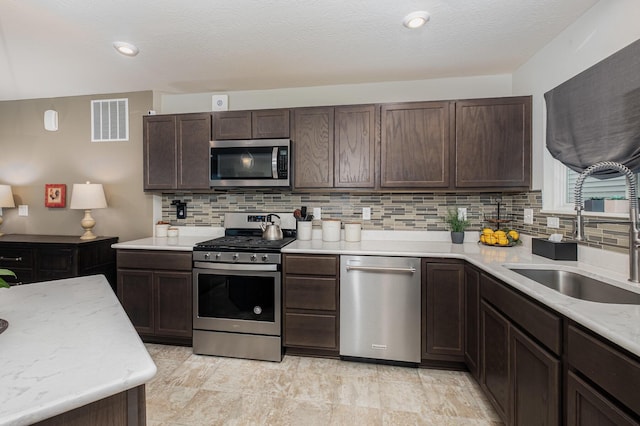 kitchen with appliances with stainless steel finishes, a sink, backsplash, dark brown cabinetry, and light stone countertops