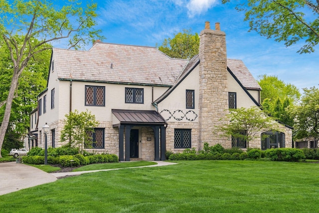 tudor-style house with stone siding, a front yard, a chimney, and a standing seam roof