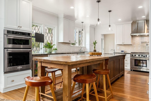 kitchen with appliances with stainless steel finishes, wall chimney exhaust hood, decorative backsplash, butcher block counters, and recessed lighting