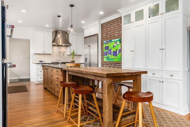 kitchen featuring stainless steel built in refrigerator, wall chimney exhaust hood, light wood-style flooring, decorative backsplash, and white cabinetry