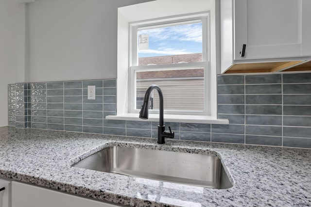 kitchen view of a sink, decorative backsplash, light stone countertops, and white cabinets