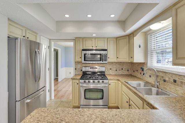 kitchen featuring stainless steel appliances, cream cabinetry, a sink, light countertops, and a wainscoted wall