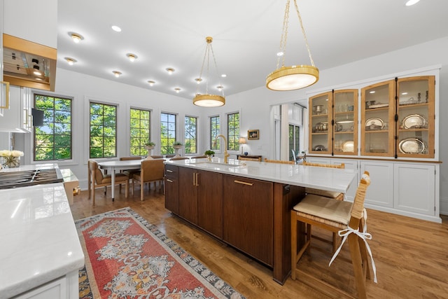 kitchen featuring a sink, a center island with sink, glass insert cabinets, light wood-type flooring, and decorative light fixtures