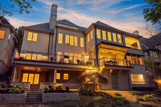 back of house at dusk featuring a balcony, ceiling fan, a chimney, stucco siding, and stone siding