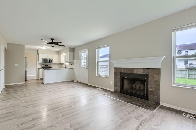 unfurnished living room with a tiled fireplace, light wood-style flooring, ceiling fan, a sink, and baseboards