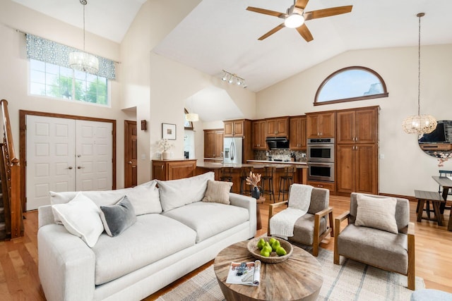 living room featuring a chandelier, high vaulted ceiling, ceiling fan, light wood-style flooring, and healthy amount of natural light