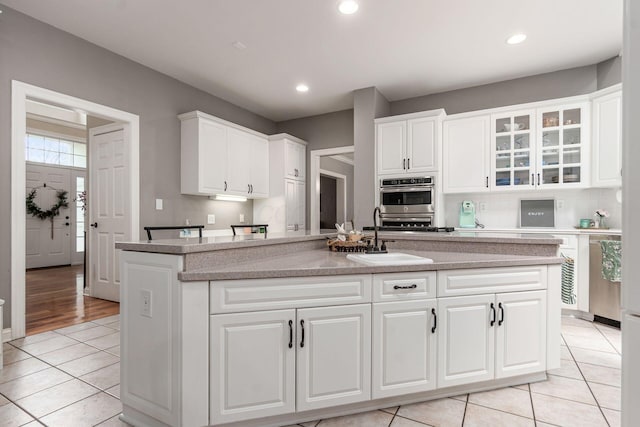 kitchen featuring a sink, light tile patterned floors, white cabinets, glass insert cabinets, and an island with sink