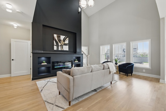 living room featuring high vaulted ceiling, light wood-style flooring, baseboards, a glass covered fireplace, and stairway