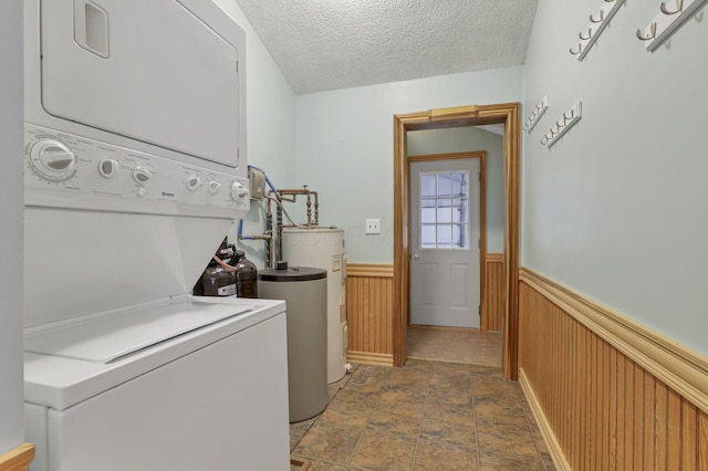 washroom with estacked washer and dryer, wainscoting, water heater, stone finish floors, and a textured ceiling