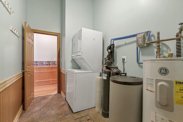 washroom with stacked washer / dryer, electric water heater, wainscoting, and stone finish flooring