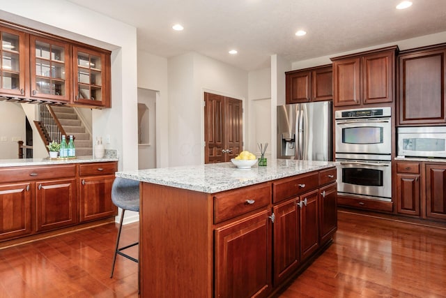 7019 Blakemore Ln, Dublin, OH 43016 - kitchen with stainless steel appliances, dark wood-style flooring, glass insert cabinets, a breakfast bar area, and light stone counters