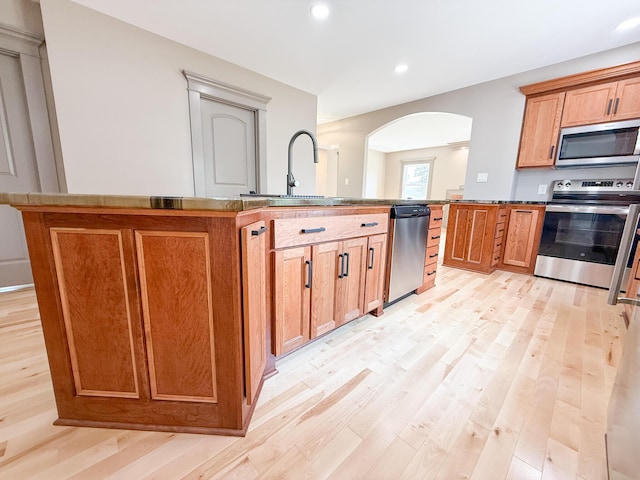 69 N Cherry St, Mount Gilead, OH 43338 - kitchen featuring appliances with stainless steel finishes, arched walkways, light wood finished floors, a kitchen island with sink, and brown cabinets