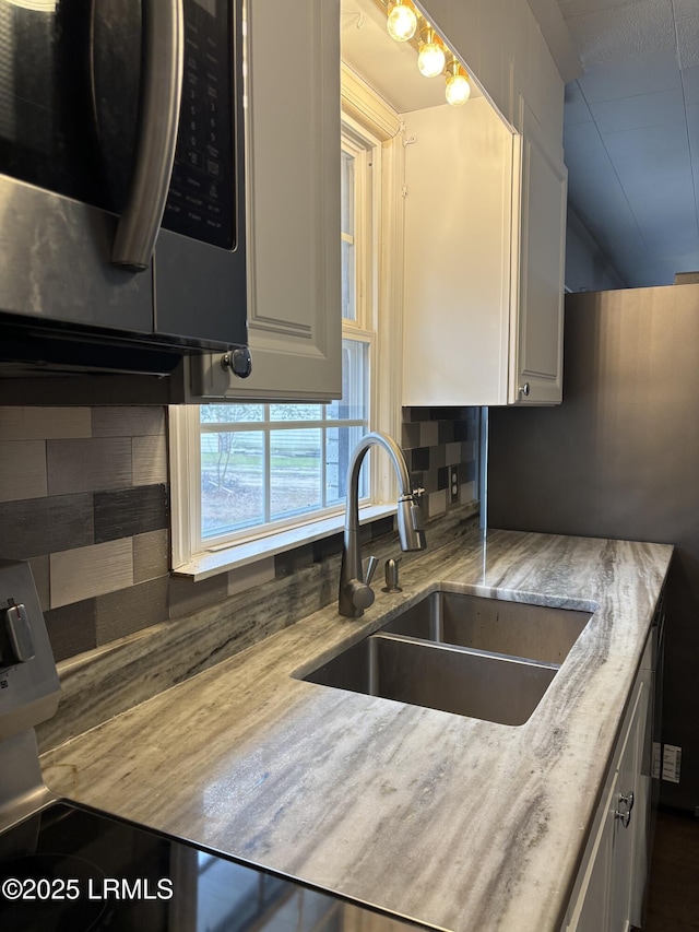 kitchen featuring stainless steel microwave, a sink, light stone countertops, white cabinetry, and backsplash