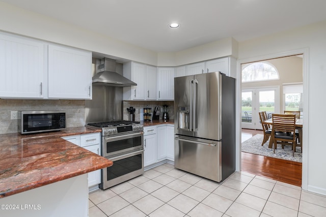 kitchen featuring tasteful backsplash, white cabinets, light tile patterned floors, stainless steel appliances, and wall chimney range hood