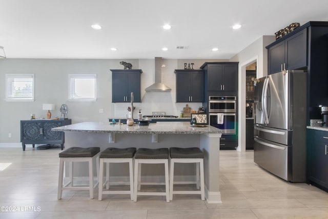 kitchen featuring a kitchen bar, stainless steel appliances, light stone countertops, a kitchen island with sink, and wall chimney range hood