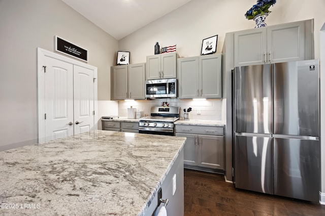 kitchen featuring appliances with stainless steel finishes, vaulted ceiling, light stone countertops, gray cabinets, and backsplash