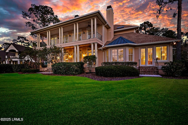 back of property at dusk featuring a ceiling fan, a lawn, a balcony, and stucco siding