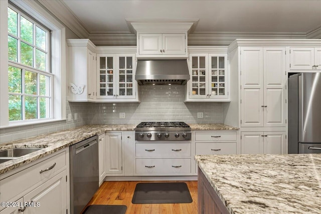 kitchen with appliances with stainless steel finishes, backsplash, ornamental molding, white cabinets, and wall chimney exhaust hood