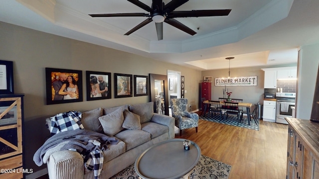 living room with ornamental molding, a raised ceiling, ceiling fan with notable chandelier, and light wood-type flooring