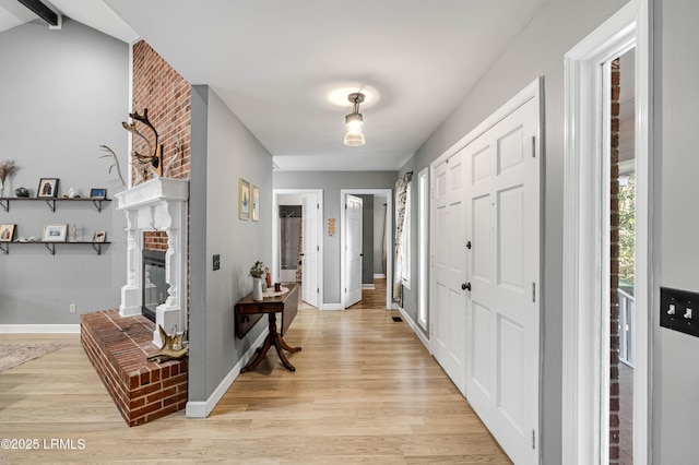 foyer entrance featuring light wood-style floors, a brick fireplace, lofted ceiling with beams, and baseboards