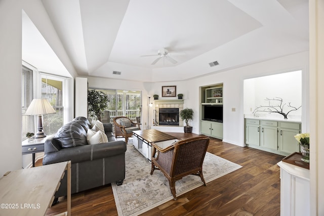 living room with built in features, ceiling fan, dark hardwood / wood-style floors, a tiled fireplace, and a raised ceiling