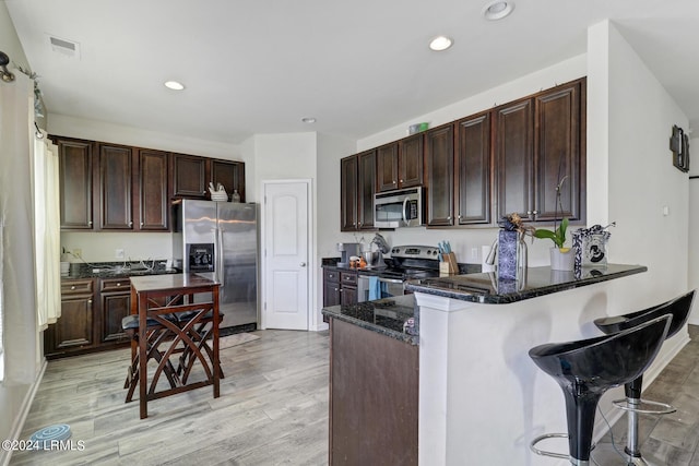 kitchen with dark brown cabinets, light hardwood / wood-style flooring, appliances with stainless steel finishes, kitchen peninsula, and dark stone counters