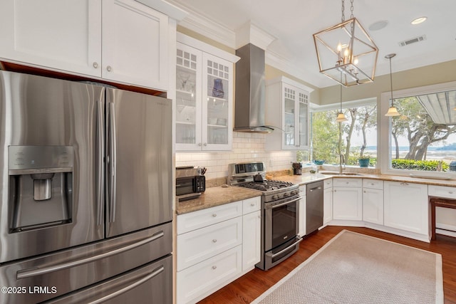 kitchen featuring glass insert cabinets, appliances with stainless steel finishes, decorative light fixtures, wall chimney range hood, and white cabinetry