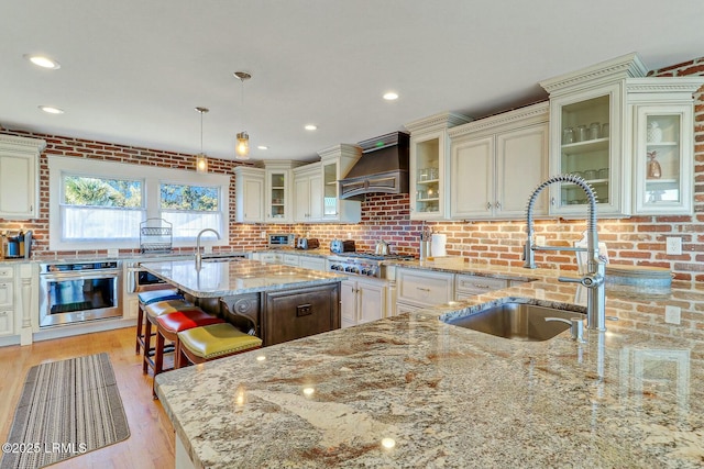 kitchen with brick wall, light stone counters, stainless steel appliances, premium range hood, and a sink