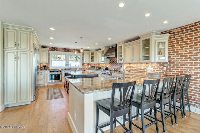 kitchen with cream cabinetry, stainless steel appliances, custom range hood, light wood-style flooring, and light stone countertops