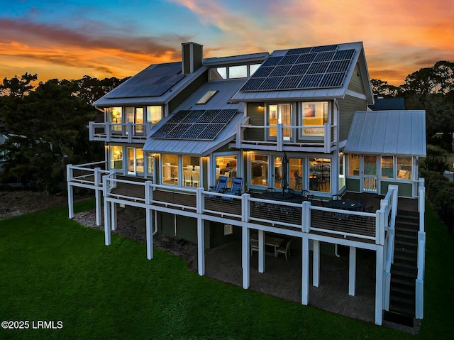 back of property at dusk with a balcony, a chimney, metal roof, a yard, and roof mounted solar panels