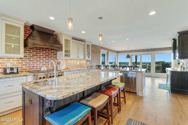 kitchen featuring a kitchen island with sink, a sink, premium range hood, dishwasher, and a peninsula