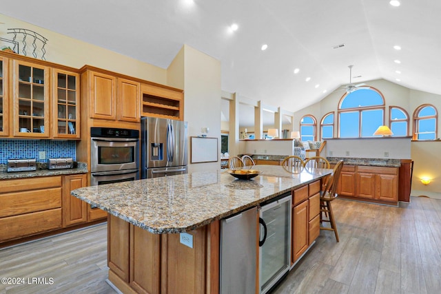 kitchen with wine cooler, light stone counters, light hardwood / wood-style flooring, appliances with stainless steel finishes, and an island with sink