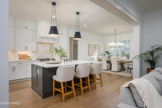 kitchen with white cabinets, backsplash, hanging light fixtures, a center island with sink, and light hardwood / wood-style flooring