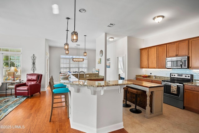 kitchen featuring brown cabinetry, decorative backsplash, appliances with stainless steel finishes, a kitchen bar, and pendant lighting