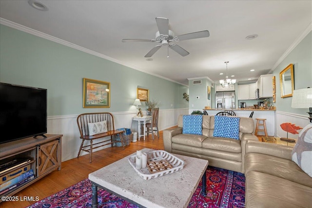 living room with ceiling fan with notable chandelier, ornamental molding, wainscoting, and wood finished floors