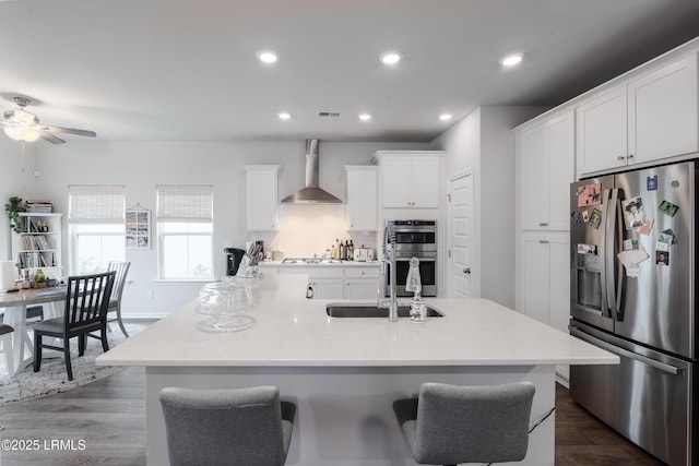 kitchen with sink, a center island with sink, appliances with stainless steel finishes, wall chimney range hood, and white cabinets