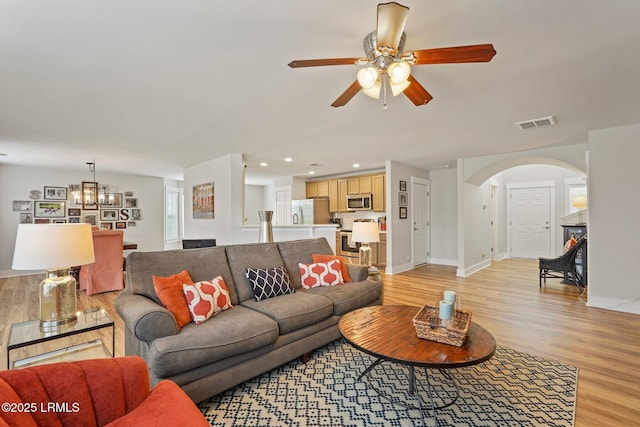living area featuring arched walkways, light wood-type flooring, ceiling fan with notable chandelier, baseboards, and visible vents