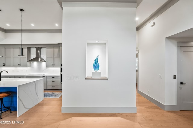 kitchen with a breakfast bar, wall chimney range hood, light wood-type flooring, light stone counters, and gray cabinetry